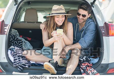 Stock Image - Couple in car boot looking at smartphone. Fotosearch