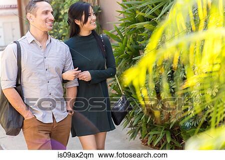 Couple in street, linking arms looking away smiling View Large Photo Image Picture - Couple in street, linking arms looking away smiling. Fotosearch
