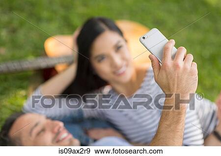 Couple lying on grass using smartphone to take selfie View Large Photo Image Stock Photo - Couple lying on grass using smartphone to take selfie. Fotosearch
