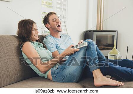 Stock Image - Couple on sofa using remote control laughing. Fotosearch