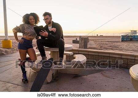 Stock Image - Couple outdoors, beside beach, looking at digital tablet, woman wearing rollerskates. Fotosearch