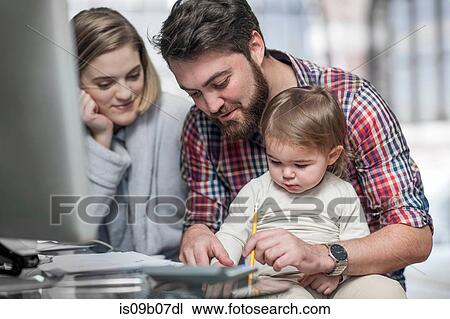 Stock Image - Couple sitting at desk, toddler sitting on man's lap watching man draw. Fotosearch