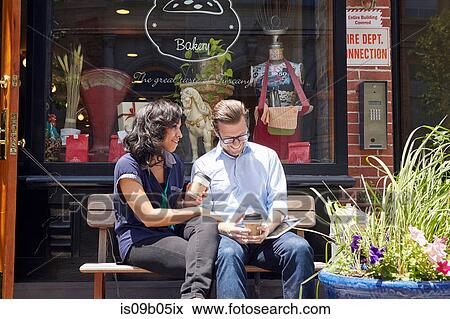 Couple sitting on bench, outside bakery, holding coffee cups View Large Photo Image Stock Photo - Couple sitting on bench, outside bakery, holding coffee cups. Fotosearch