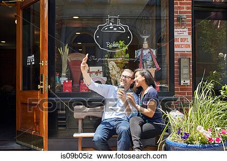 Couple sitting on bench, outside bakery, holding coffee cups, taking selfie with smartphone View Large Photo Image Stock Photo - Couple sitting on bench, outside bakery, holding coffee cups, taking selfie with smartphone. Fotosearch