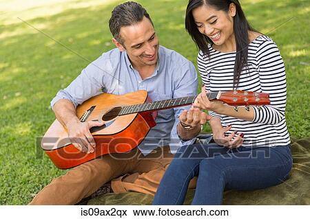 Stock Photo - Couple sitting on grass playing acoustic guitar smiling. Fotosearch