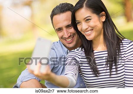 Couple sitting on grass using smartphone to take selfie View Large Photo Image Stock Photo - Couple sitting on grass using smartphone to take selfie. Fotosearch
