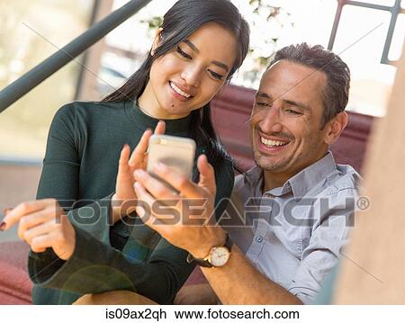 Stock Image - Couple sitting on stairway using smartphone smiling. Fotosearch