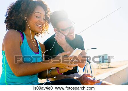 Couple sitting on wall, sharing earphones, listening to music, using smartphone View Large Photo Image Stock Image - Couple sitting on wall, sharing earphones, listening to music, using smartphone. Fotosearch