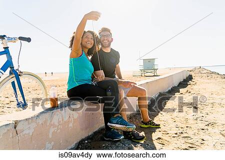 Couple sitting on wall taking self portrait using smartphone View Large Photo Image Stock Photo - Couple sitting on wall taking self portrait using smartphone. Fotosearch