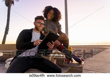 Couple sitting outdoors, beside beach, looking at digital tablet, woman wearing rollerskates View Large Photo Image Stock Image - Couple sitting outdoors, beside beach, looking at digital tablet, woman wearing rollerskates. Fotosearch