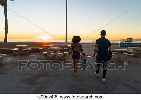 Stock Photograph - Couple walking near beach, holding skateboards, rear view. Fotosearch