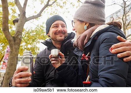 Stock Image - Couple wearing knit hats holding drinks flask in park. Fotosearch