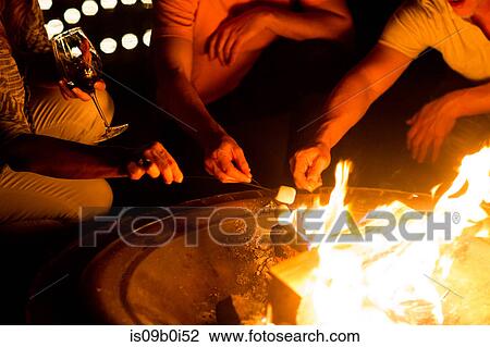Stock Image - Cropped shot of mature adult couples toasting marshmallows on patio fire at night. Fotosearch