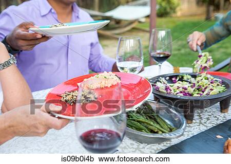 Stock Image - Cropped shot of men serving food onto plates at garden table. Fotosearch