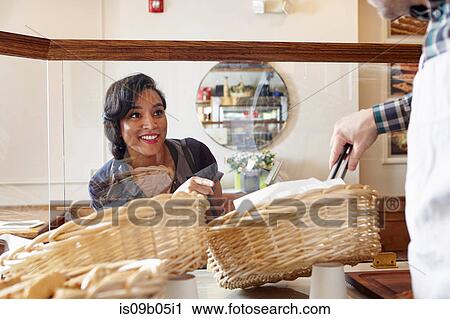Stock Image - Customer in bakery pointing to baked goods, worker selecting it for her with tongs. Fotosearch