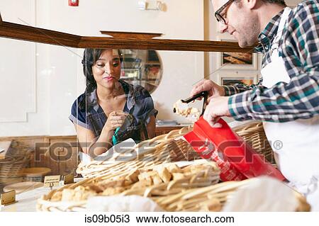 Stock Image - Customer in bakery pointing to baked goods, worker placing goods in bag. Fotosearch