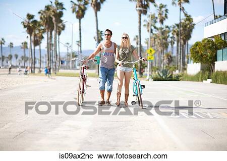 Stock Image - Cycling couple strolling with cycles at Venice Beach, Los Angeles, California, USA. Fotosearch