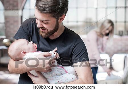 Father holding baby daughter, mother taking telephone call in background View Large Photo Image Stock Photograph - Father holding baby daughter, mother taking telephone call in background. Fotosearch