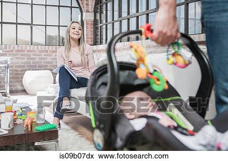 Stock Photo - Father leaving house with baby girl in car seat, mother sitting on sofa, working. Fotosearch