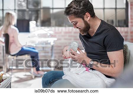Father sitting, feeding baby daughter, mother sitting at desk, working, behind View Large Photo Image Stock Photograph - Father sitting, feeding baby daughter, mother sitting at desk, working, behind. Fotosearch