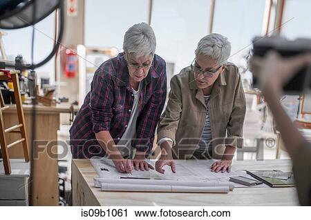 Stock Image - Female carpenters discussing blueprint in furniture making workshop. Fotosearch