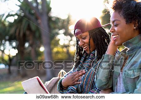 Stock Image - Female friends relaxing in park, sharing earphones. Fotosearch