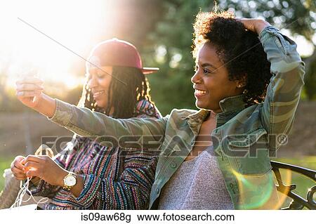 Stock Image - Female friends relaxing in park, woman taking self portrait using smartphone. Fotosearch