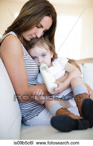 Stock Image - Female toddler on mother's lap drinking from baby bottle. Fotosearch