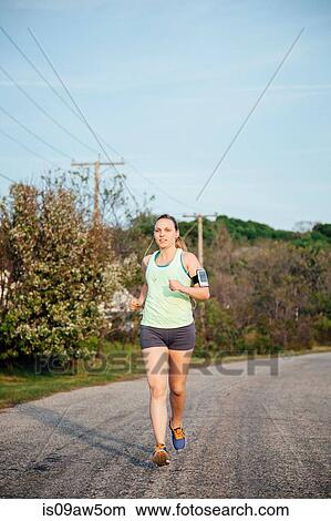 Picture - Full length front of woman wearing activity tracker jogging on road. Fotosearch