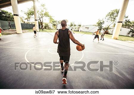 Stock Photo - Full length rear view of young man walking on basketball court holding basketball. Fotosearch