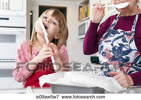 Stock Photo - Girl and grandmother licking cake mix from spatula's at kitchen counter. Fotosearch
