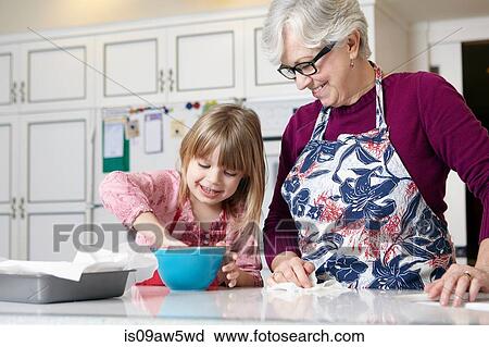 Stock Image - Girl and grandmother mixing cake in bowl at kitchen counter. Fotosearch