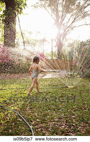 Stock Photo - Girl in bathing costume playing in sunlit garden sprinkler water. Fotosearch