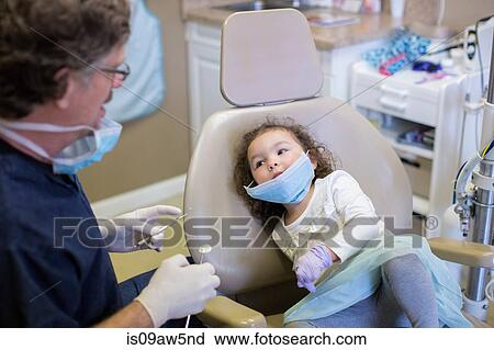 Stock Image - Girl in dentist chair watching dentist prepare dental equipment. Fotosearch