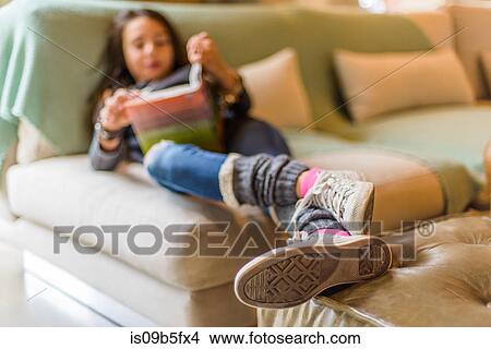 Picture - Girl lying on sofa, legs raised on coffee table reading book. Fotosearch