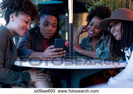 Stock Image - Group of female friends sitting at table, looking at smartphone, outdoors. Fotosearch