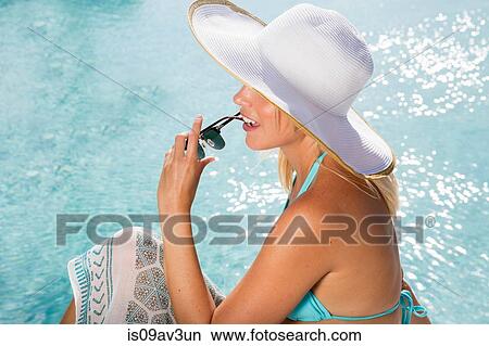 High angle side view of young woman wearing sunhat sitting by swimming pool holding sunglasses View Large Photo Image Picture - High angle side view of young woman wearing sunhat sitting by swimming pool holding sunglasses. Fotosearch