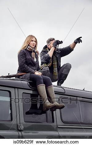 Stock Image - Hunting couple, on top of off road vehicle with shot gun and binoculars. Fotosearch