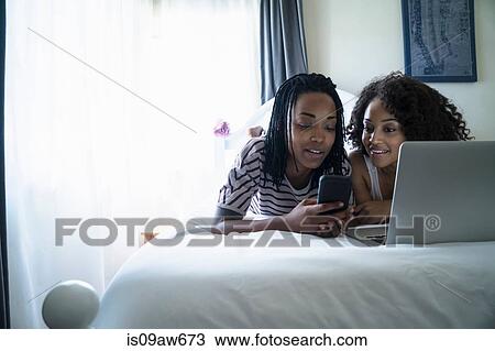 Stock Image - Lesbian couple lying on bed, using laptop. Fotosearch