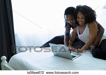 Stock Image - Lesbian couple sitting on bed, looking at laptop. Fotosearch