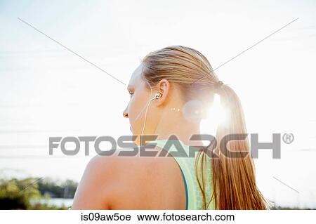 Stock Image - Low angle rear view of woman with ponytail wearing earbuds looking away. Fotosearch