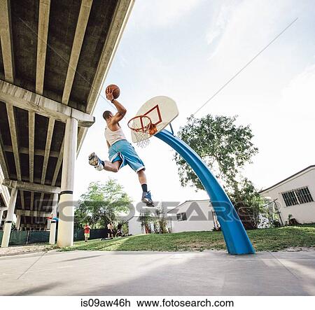 Stock Image - Low angle view of young man in mid air holding basketball jumping for hoop. Fotosearch