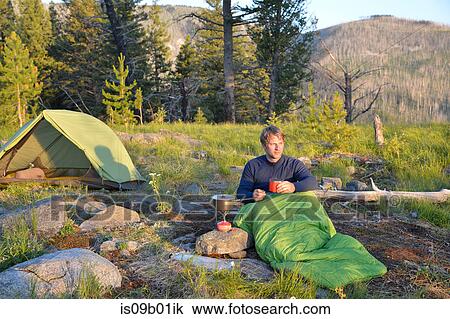 Stock Image - Male camper in sleeping bag prepares a meal on camping stove at Midnight Ridge, Colville National Forest, Washington State, USA. Fotosearch