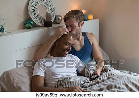 Stock Photography - Male couple lying in bed together, looking at smartphone. Fotosearch