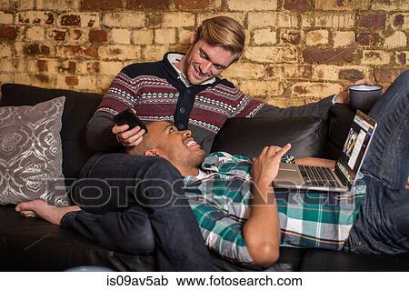 Stock Image - Male couple relaxing on sofa together, looking at laptop. Fotosearch