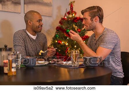Stock Image - Male couple sitting at table, having breakfast, Christmas tree in background. Fotosearch
