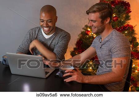 Stock Photograph - Male couple sitting at table, looking at laptop, smiling. Fotosearch