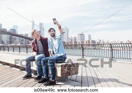 Stock Image - Male couple sitting on riverside by Brooklyn Bridge taking smartphone selfie, New York, USA. Fotosearch