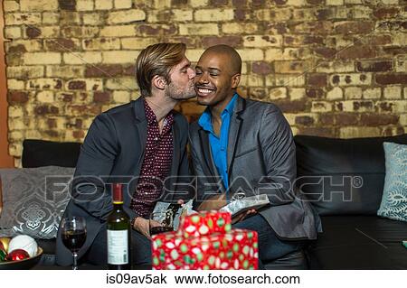 Stock Image - Male couple sitting on sofa, exchanging Christmas gifts, young man kissing partner on cheek. Fotosearch