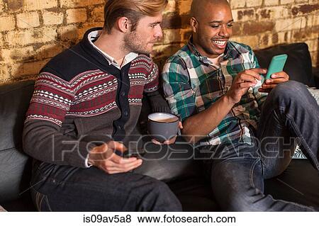 Stock Photo - Male couple sitting on sofa, looking at smartphones. Fotosearch